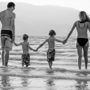 dad, two sons, and wife holding hands while wearing swimsuits in shallow water on a beach